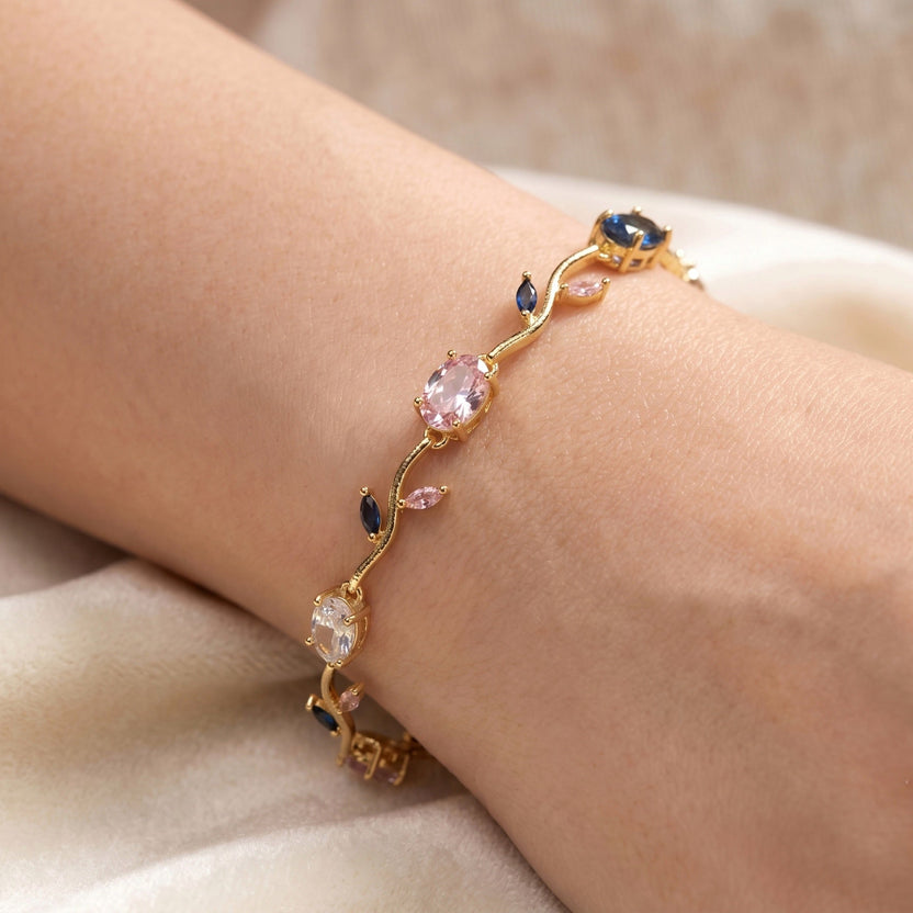 A gold charm bracelet with eye and crescent moon charms worn on a woman's wrist, resting on a light beige stone slab with linen fabric and a small dish in the background.