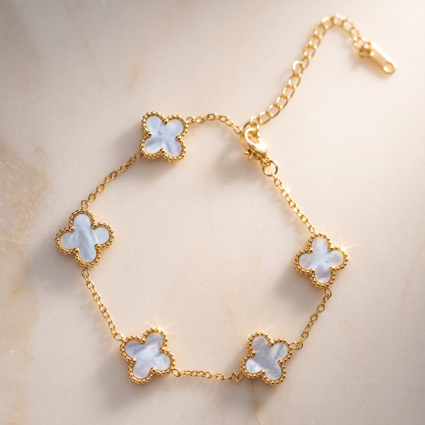 A gold chain bracelet with five white mother-of-pearl four-leaf clover motifs, displayed on a light beige marble surface.