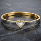 A gold bangle bracelet with a textured dark gray inlay and a central heart-shaped mother-of-pearl centerpiece surrounded by small clear stones, displayed on a dark marble surface.