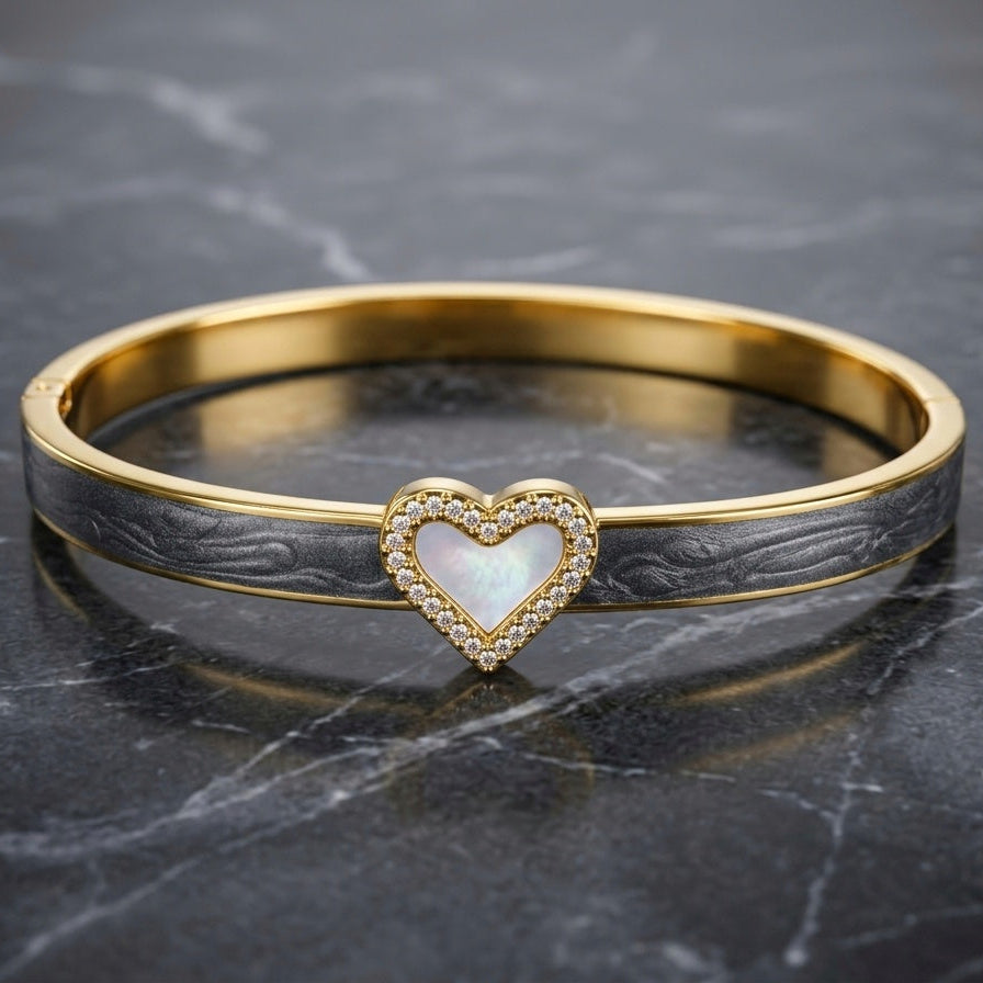 A gold bangle bracelet with a textured dark gray inlay and a central heart-shaped mother-of-pearl centerpiece surrounded by small clear stones, displayed on a dark marble surface.