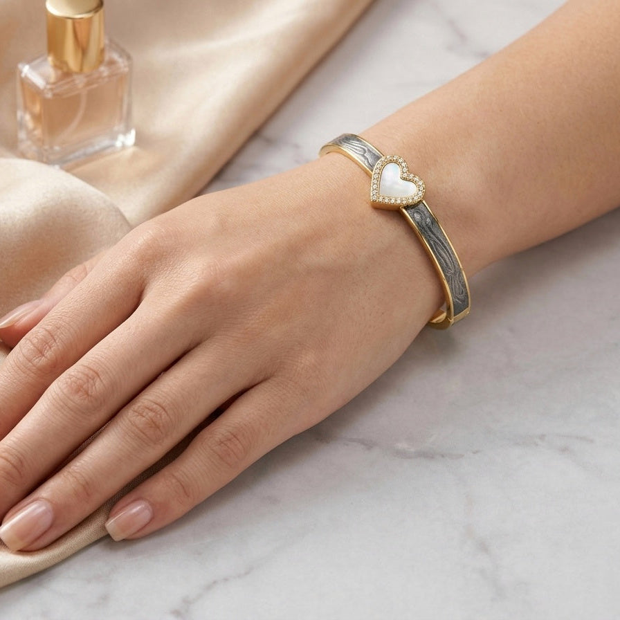 A gold-toned cuff bracelet with a white heart-shaped inlay, worn on a woman's wrist. The hand rests on draped beige satin fabric next to a perfume bottle on a light marble surface.