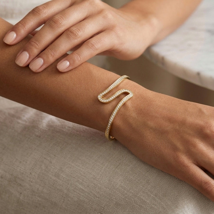 A close-up lifestyle shot of a woman's wrist wearing a gold open cuff bracelet paved with small clear stones, with her other hand resting nearby and a marble table in the background.