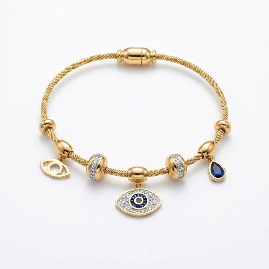A gold cable chain bracelet with multiple charms, including an evil eye pendant, a teardrop charm with a dark blue stone, and two bezel-set clear crystal charms, displayed on a clean white background.