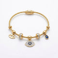 A gold cable chain bracelet with multiple charms, including an evil eye pendant, a teardrop charm with a dark blue stone, and two bezel-set clear crystal charms, displayed on a clean white background.