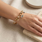 A gold charm bracelet with eye and crescent moon charms worn on a woman's wrist, resting on a light beige stone slab with linen fabric and a small dish in the background.