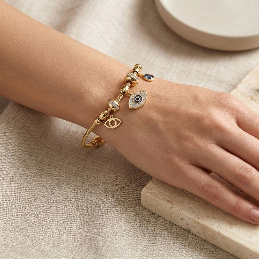 A gold charm bracelet with eye and crescent moon charms worn on a woman's wrist, resting on a light beige stone slab with linen fabric and a small dish in the background.