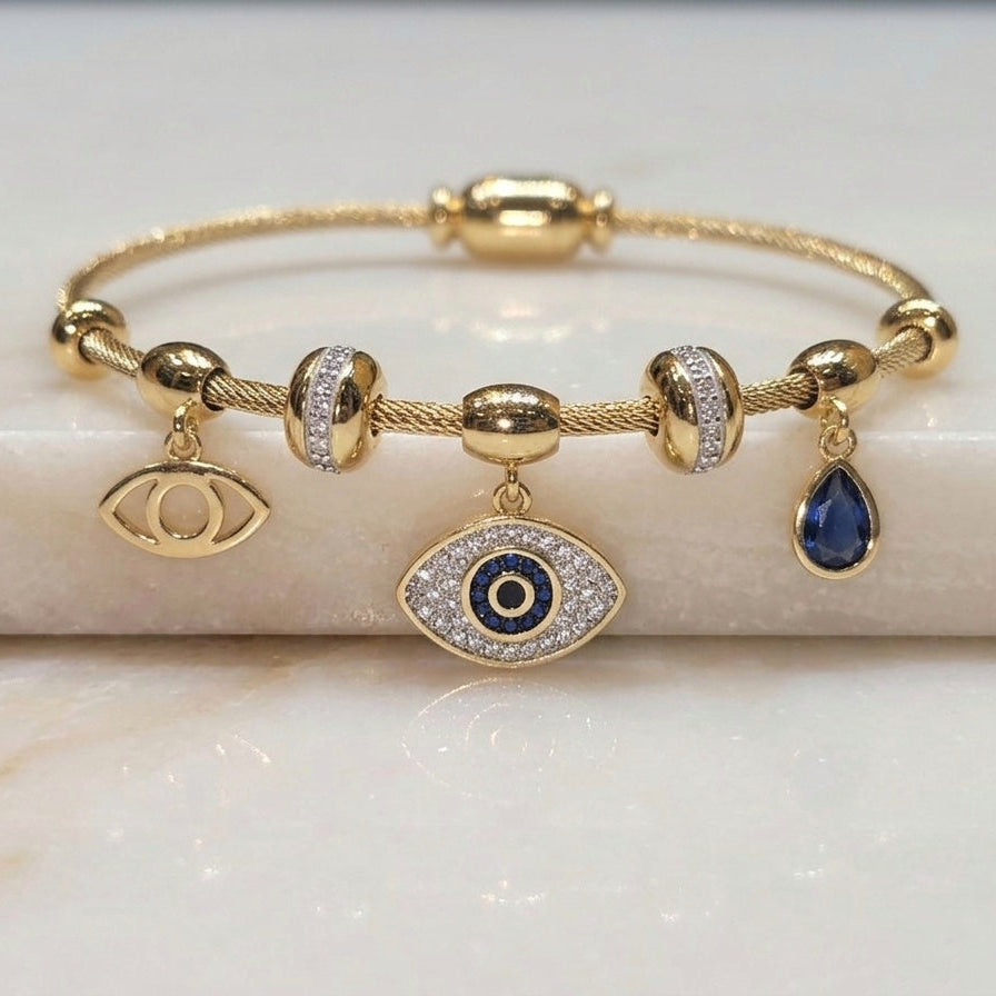 A gold bangle bracelet with evil eye charms, displayed on a white marble block. The bracelet features a gold chain with cylindrical and round beads, some adorned with clear pavé stones, and three dangling charms: a gold eye, a larger gold eye with a blue and white center, and a teardrop-shaped dark blue stone.