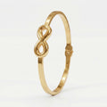 A polished gold bangle bracelet featuring a central infinity symbol, set against a clean white background.