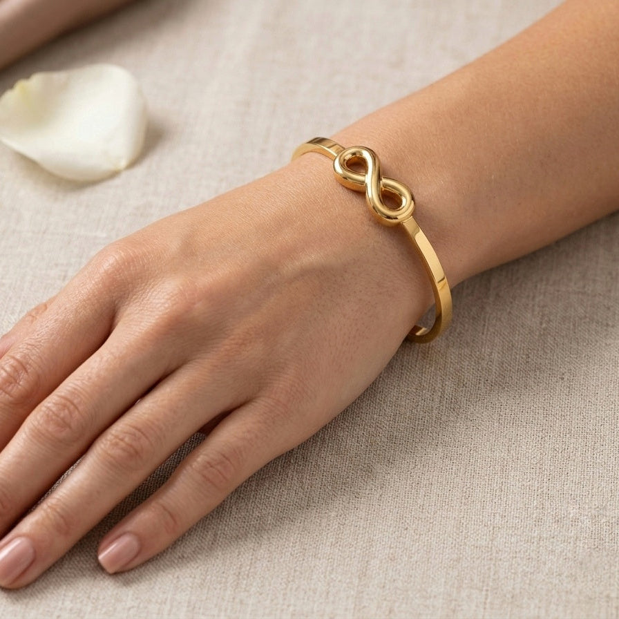 A gold infinity bracelet worn on a woman's wrist, shown in a close-up lifestyle shot against a neutral beige textured fabric background.