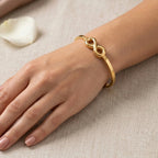 A gold infinity bracelet worn on a woman's wrist, shown in a close-up lifestyle shot against a neutral beige textured fabric background.