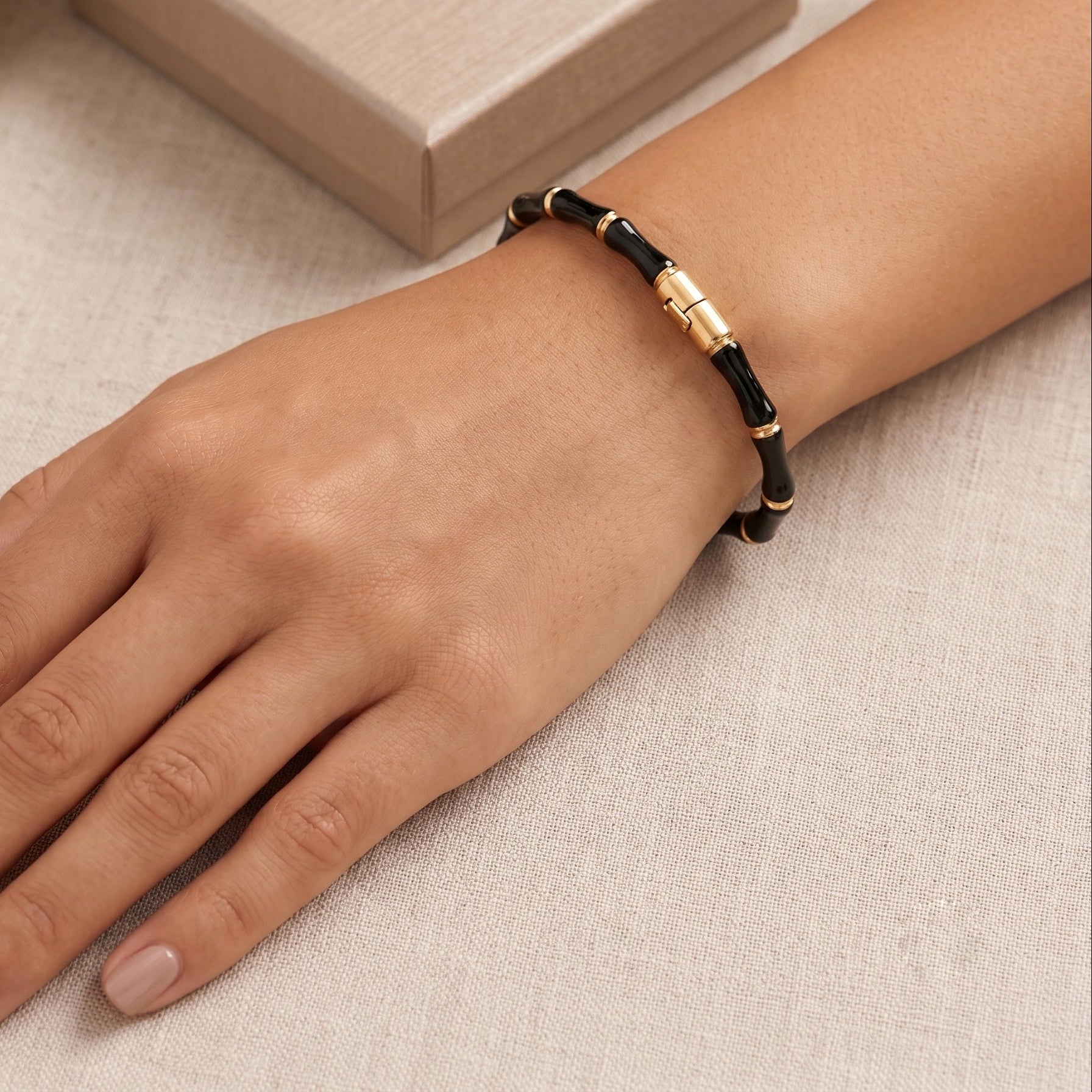 A close-up of a person's hand and wrist wearing a black beaded bracelet with a polished gold clasp, resting on a light beige textured fabric surface.