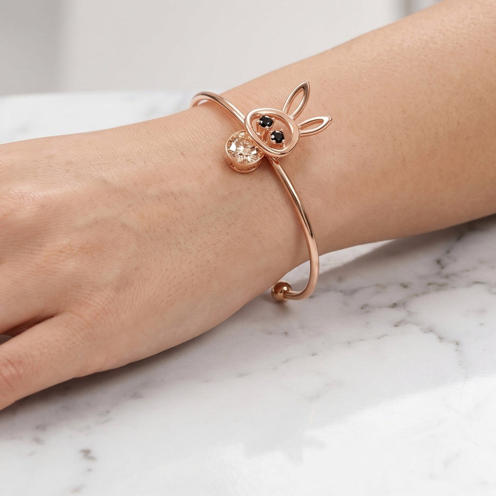 A close-up of a person's hand resting on a white marble surface, wearing a rose gold open cuff bracelet with a bunny head charm and a matching rose gold ring.