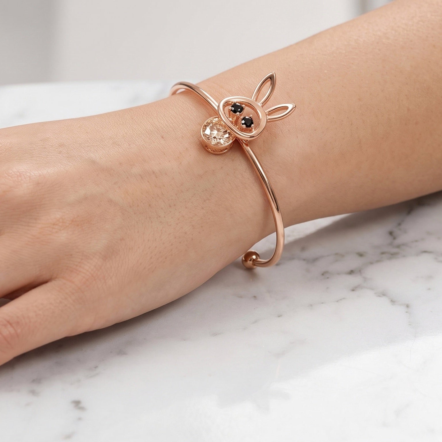 A close-up of a person's hand resting on a white marble surface, wearing a rose gold open cuff bracelet with a bunny head charm and a matching rose gold ring.