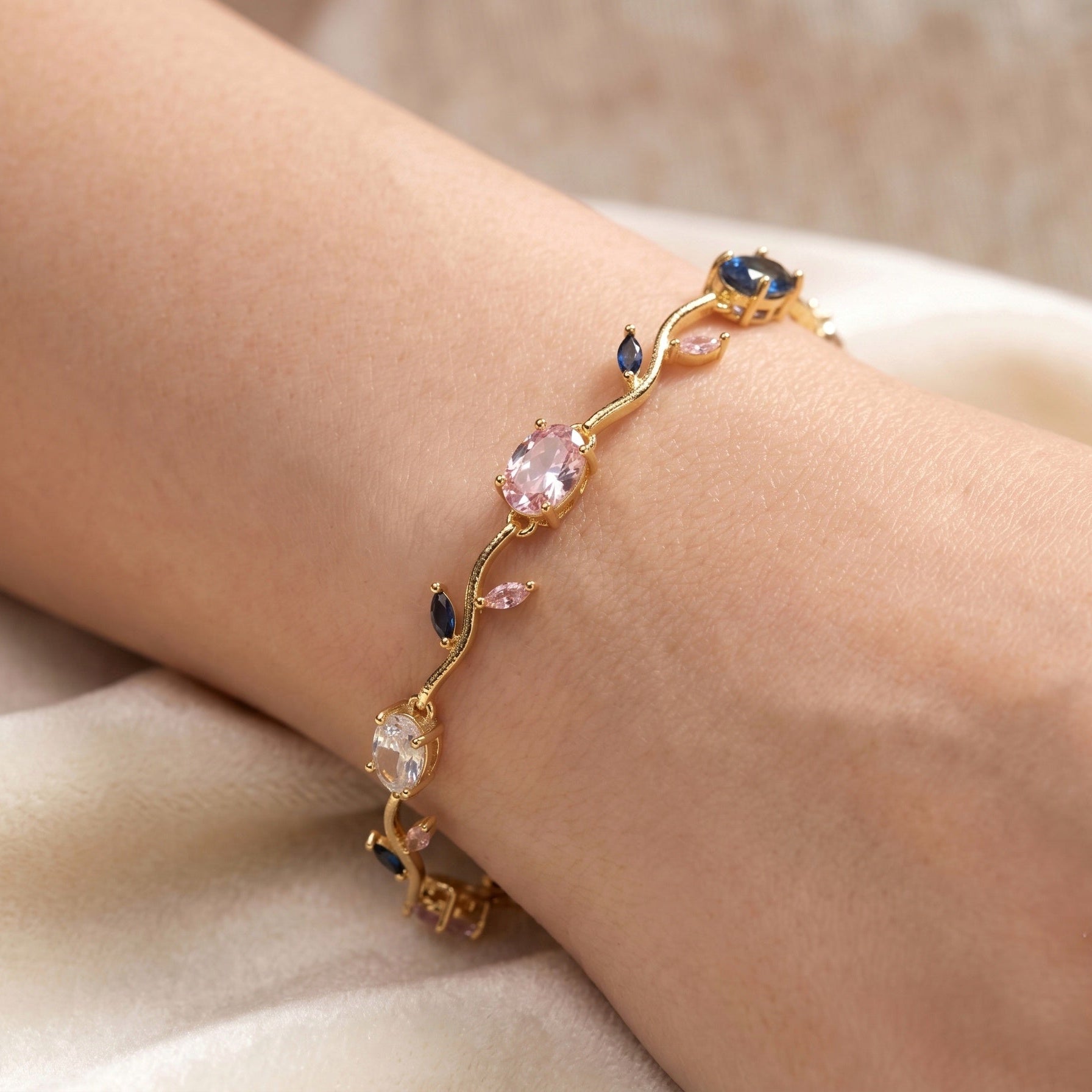A delicate gold bracelet with pink, dark blue, and clear gemstones, shown worn on a person's wrist against a soft beige fabric background.