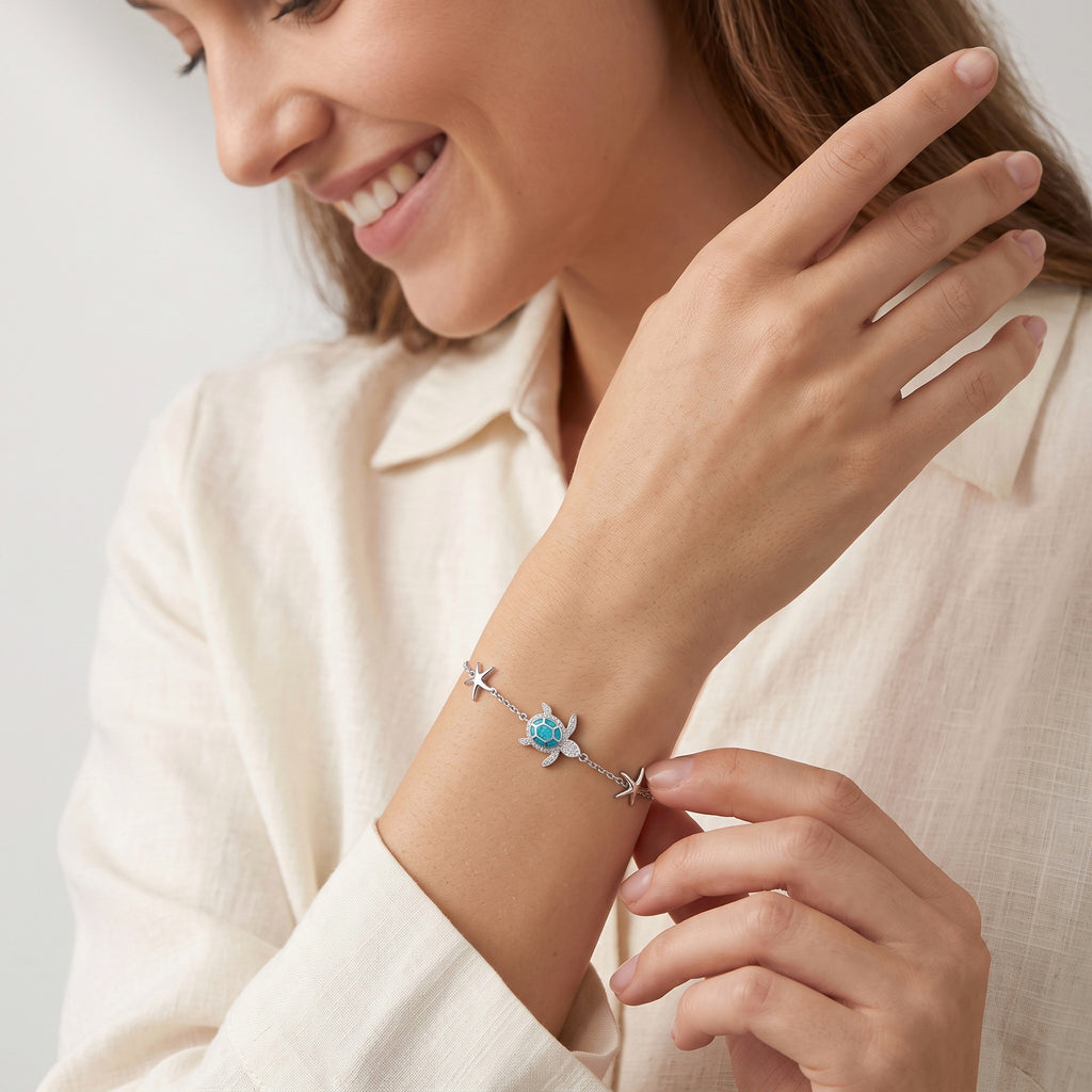 A close-up lifestyle shot of a woman wearing a delicate silver bracelet on her wrist. The bracelet features a small blue sea turtle charm flanked by two silver starfish.