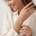 A close-up lifestyle shot of a woman wearing a delicate silver bracelet on her wrist. The bracelet features a small blue sea turtle charm flanked by two silver starfish.
