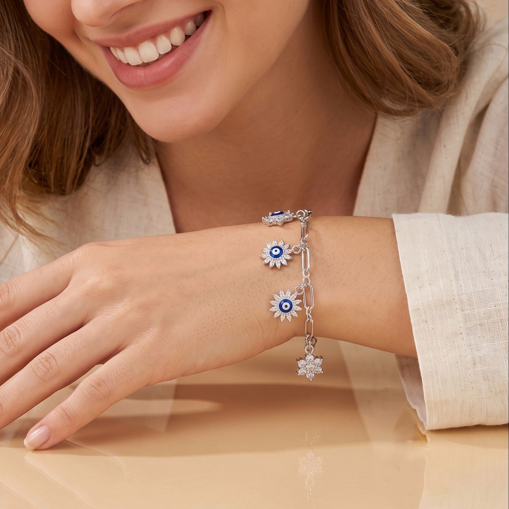 A close-up of a woman's hand and wrist wearing a silver charm bracelet with blue and white evil eye flower charms, set against a soft beige background.