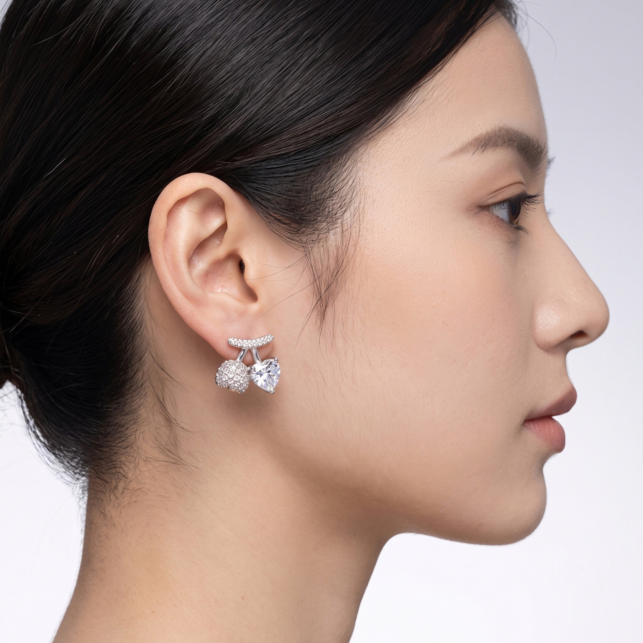 A pair of silver-toned stud earrings featuring a central heart-shaped clear gemstone, flanked by round pavé-set clear stones and a curved pavé top bar, 