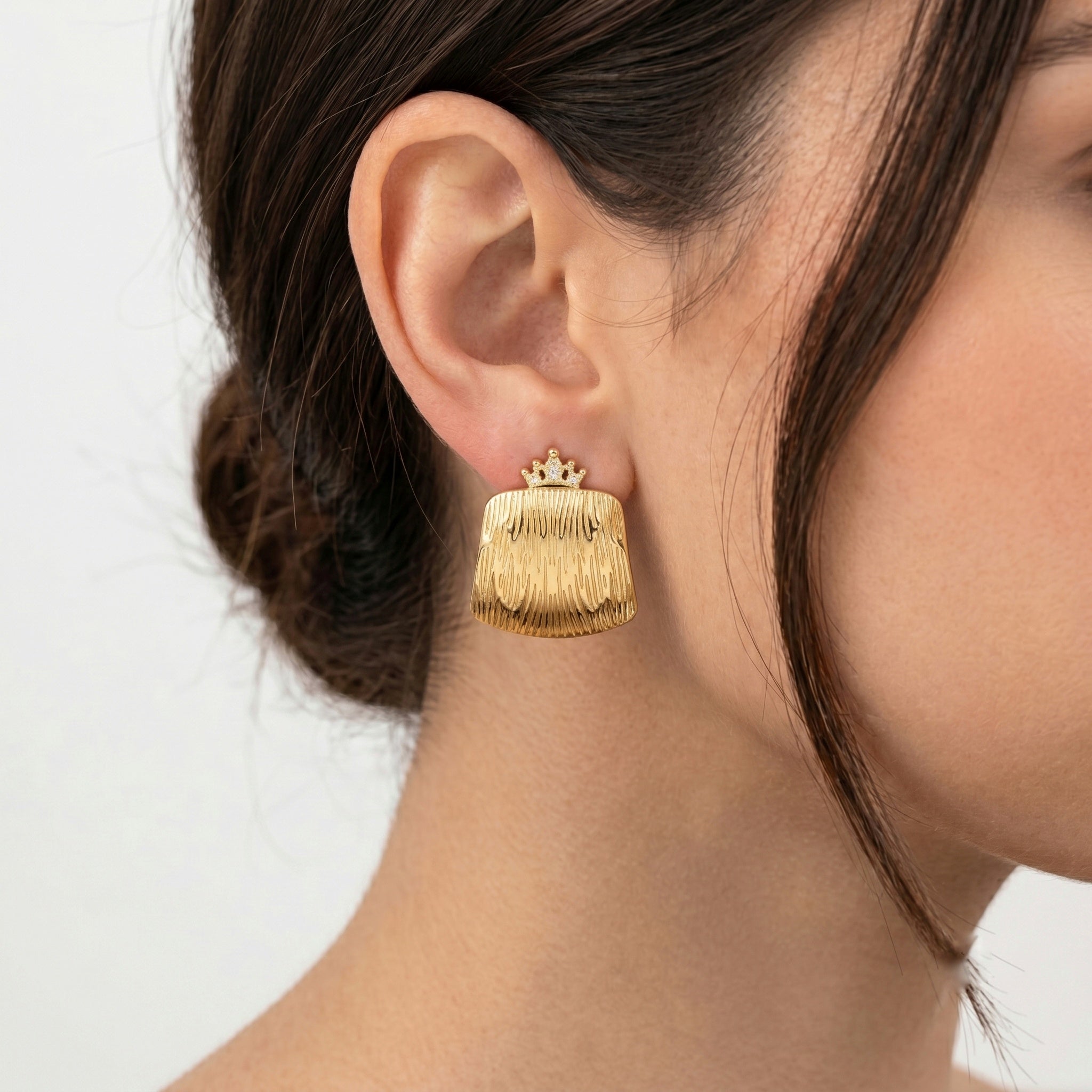 A close-up of a model's ear wearing a A pair of gold clutch-style earrings with a crown-shaped top and a vertically ribbed, textured body