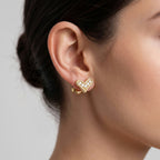model wearing a small gold heart-shaped huggie hoop earring adorned with tiny clear stones.