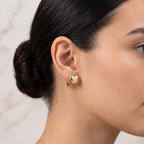 A close-up of a woman's ear wearing a small gold heart-shaped stud earring with a tiny clear gemstone, against a light marble background.
