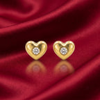 A pair of gold heart-shaped stud earrings, each featuring a round clear gemstone in the center, displayed on a draped red satin fabric background.