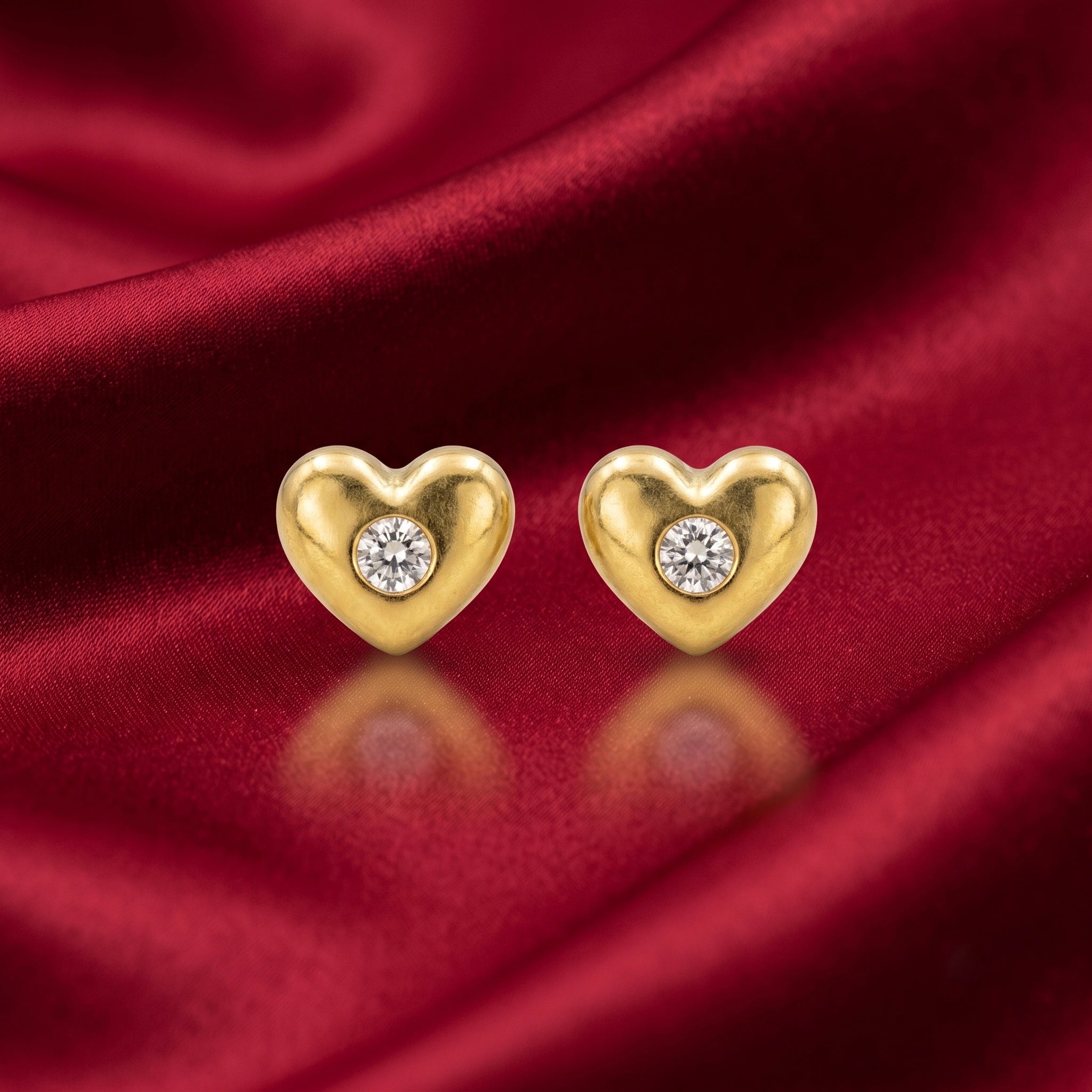 A pair of gold heart-shaped stud earrings, each featuring a round clear gemstone in the center, displayed on a draped red satin fabric background.