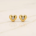 A pair of gold heart-shaped stud earrings, each featuring a small round clear gemstone in the center, displayed on a light beige marble surface.