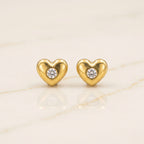 A pair of gold heart-shaped stud earrings, each featuring a small round clear gemstone in the center, displayed on a light beige marble surface.
