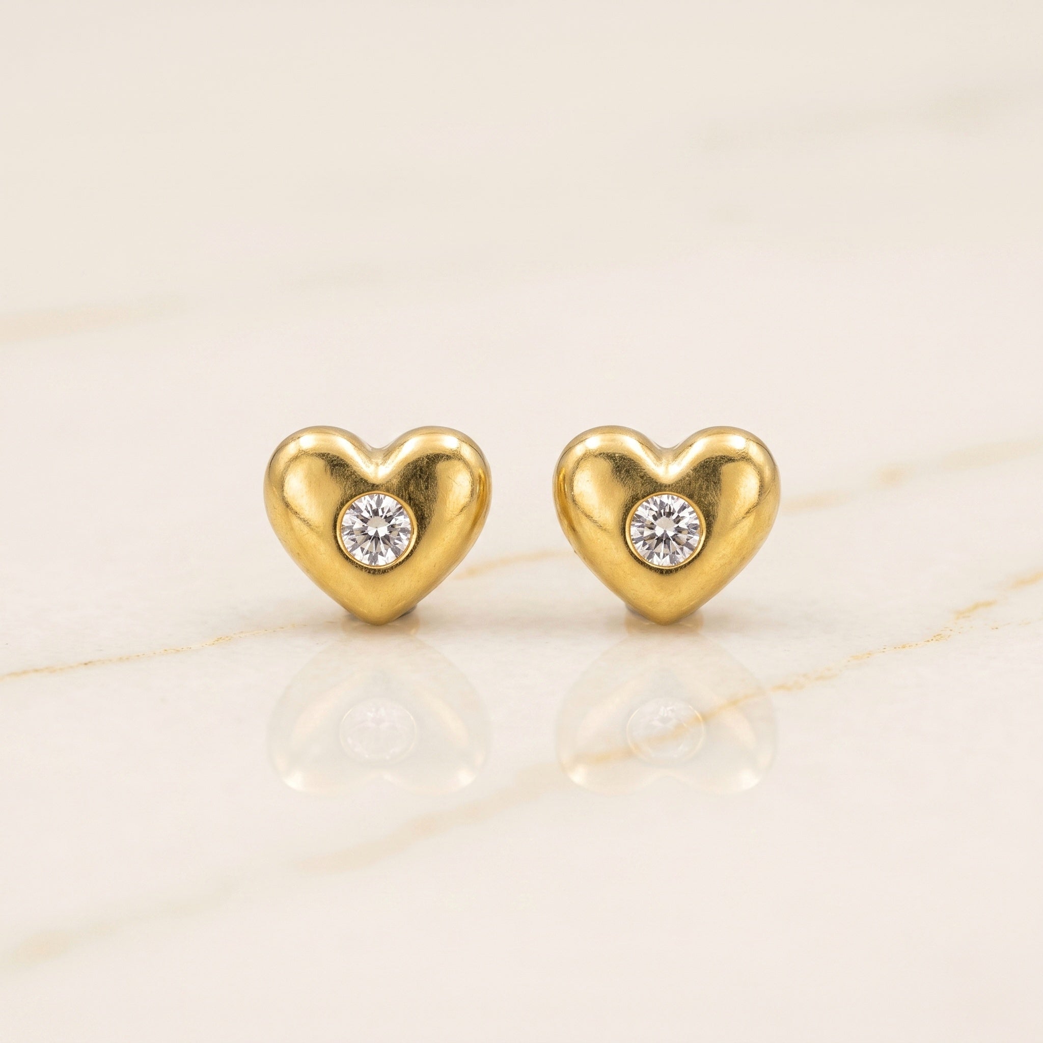 A pair of gold heart-shaped stud earrings, each featuring a small round clear gemstone in the center, displayed on a light beige marble surface.