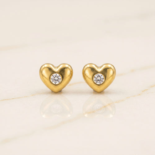 A pair of gold heart-shaped stud earrings, each featuring a small round clear gemstone in the center, displayed on a light beige marble surface.