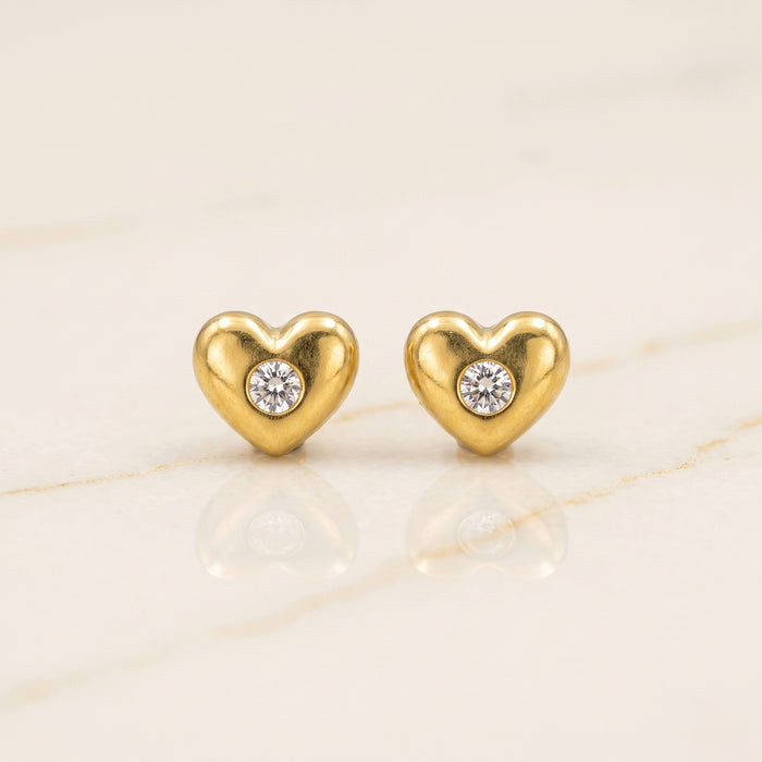 A pair of gold heart-shaped stud earrings, each featuring a small round clear gemstone in the center, displayed on a light beige marble surface.