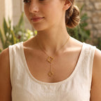 A woman wearing a delicate gold lariat necklace with two small clover-shaped pendants, styled with a white linen tank top against a softly blurred outdoor background.