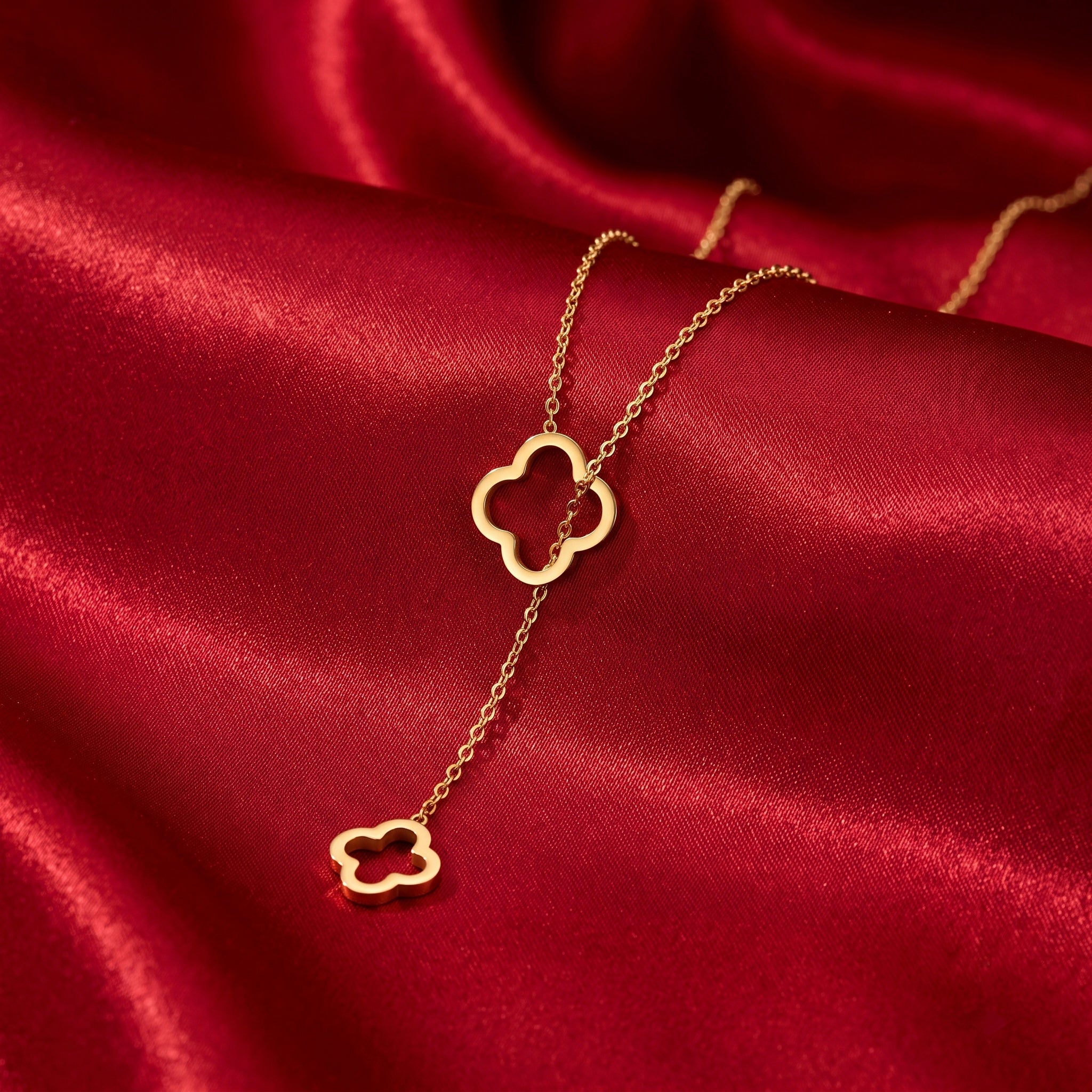 A delicate gold necklace with two interlocking clover-shaped pendants, displayed on a rich, draped red satin fabric.