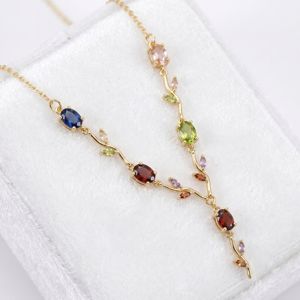 A gold vine necklace with multiple colorful gemstones, displayed on a white textured jewelry box. The necklace features a gold chain with branches and leaves set with oval and round-cut gemstones in shades of blue, red, green, pink, and purple.