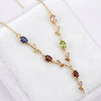 A gold vine necklace with multiple colorful gemstones, displayed on a white textured jewelry box. The necklace features a gold chain with branches and leaves set with oval and round-cut gemstones in shades of blue, red, green, pink, and purple.