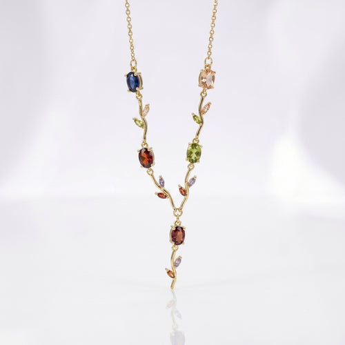 A delicate gold necklace featuring a vine-like pendant with multiple colorful gemstones in blue, green, red, and pink, displayed on a clean white background.