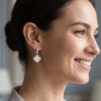 A close-up side profile of a woman wearing a delicate silver hoop earring with a white flower-shaped drop, set against a softly blurred indoor background.