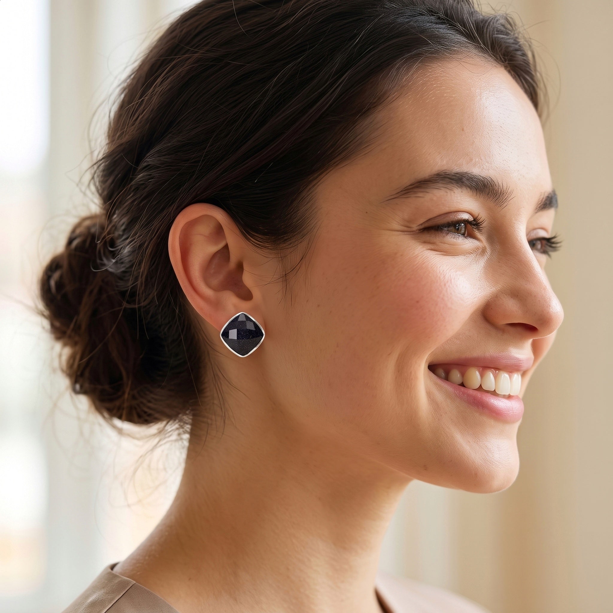 A woman with her hair in a low bun is shown in profile, wearing a small geometric stud earring with a dark blue-gray center and a silver-toned border.