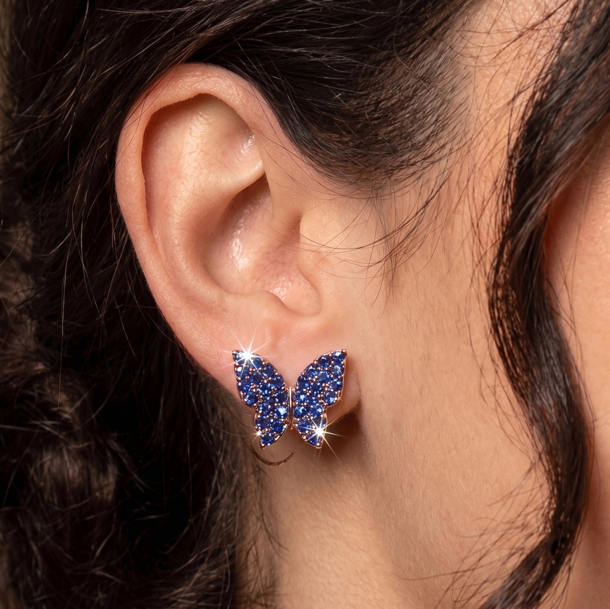 A close-up of a person's ear wearing a small butterfly-shaped stud earring covered in sparkling deep blue stones with silver-toned metal outlines.
