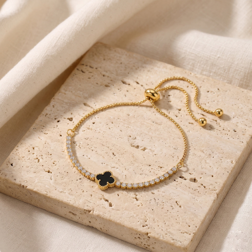 A delicate gold-tone adjustable bracelet with a black clover charm and a row of small clear stones, displayed on a beige stone slab with draped fabric.