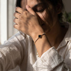 A close-up lifestyle shot of a woman wearing a delicate gold chain bracelet with a black four-leaf clover charm and small dangling gold beads, set against a softly lit, warm-toned background.