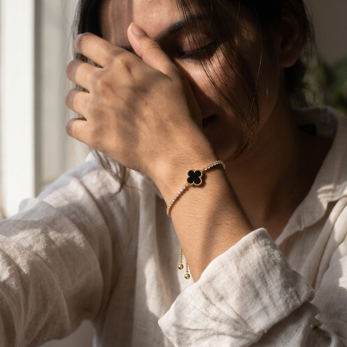 A close-up lifestyle shot of a woman wearing a delicate gold chain bracelet with a black four-leaf clover charm and small dangling gold beads, set against a softly lit, warm-toned background.
