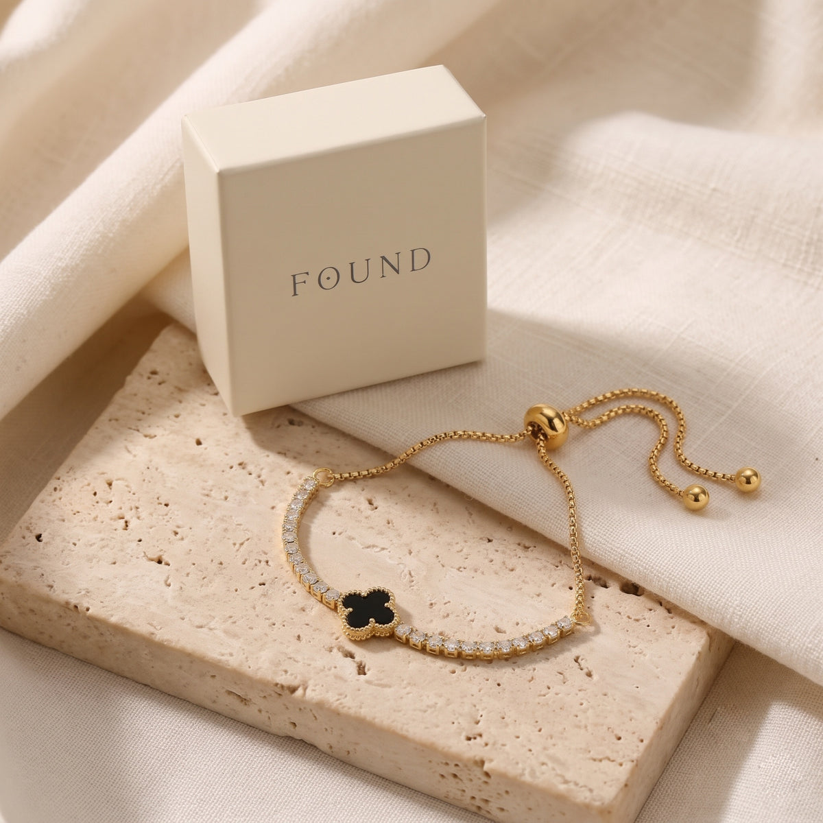 A gold-tone adjustable bracelet with a black clover charm and a row of clear stones, displayed next to a cream-colored 'FOUND' jewelry box on a textured stone slab.