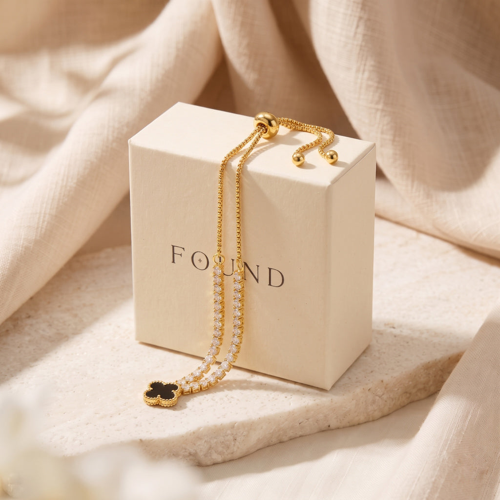 A gold-toned double-strand chain bracelet with a black clover pendant and small pearl-like accents, draped over a cream-colored jewelry box labeled 'FOUND', set against a soft beige fabric backdrop.