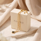 A gold-toned double-strand chain bracelet with a black clover pendant and small pearl-like accents, draped over a cream-colored jewelry box labeled 'FOUND', set against a soft beige fabric backdrop.