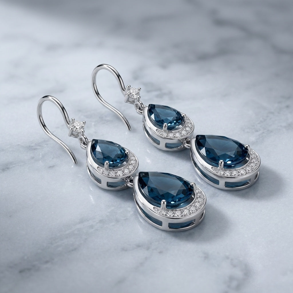 A pair of silver-tone drop earrings with teardrop-shaped dark blue gemstones and small clear accent stones, displayed on a light marble surface.