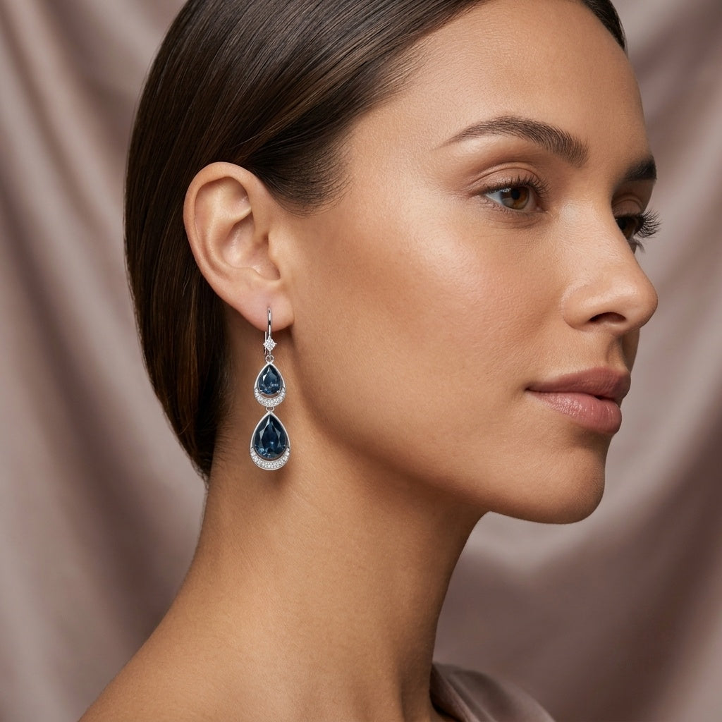 A close-up side profile of a woman wearing a single teardrop-shaped drop earring with deep blue gemstones and silver-toned settings, against a soft draped taupe background.