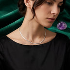 A close-up of a woman wearing a delicate silver snake chain necklace, styled with a black top and a draped dark green velvet background with a purple crystal accent.