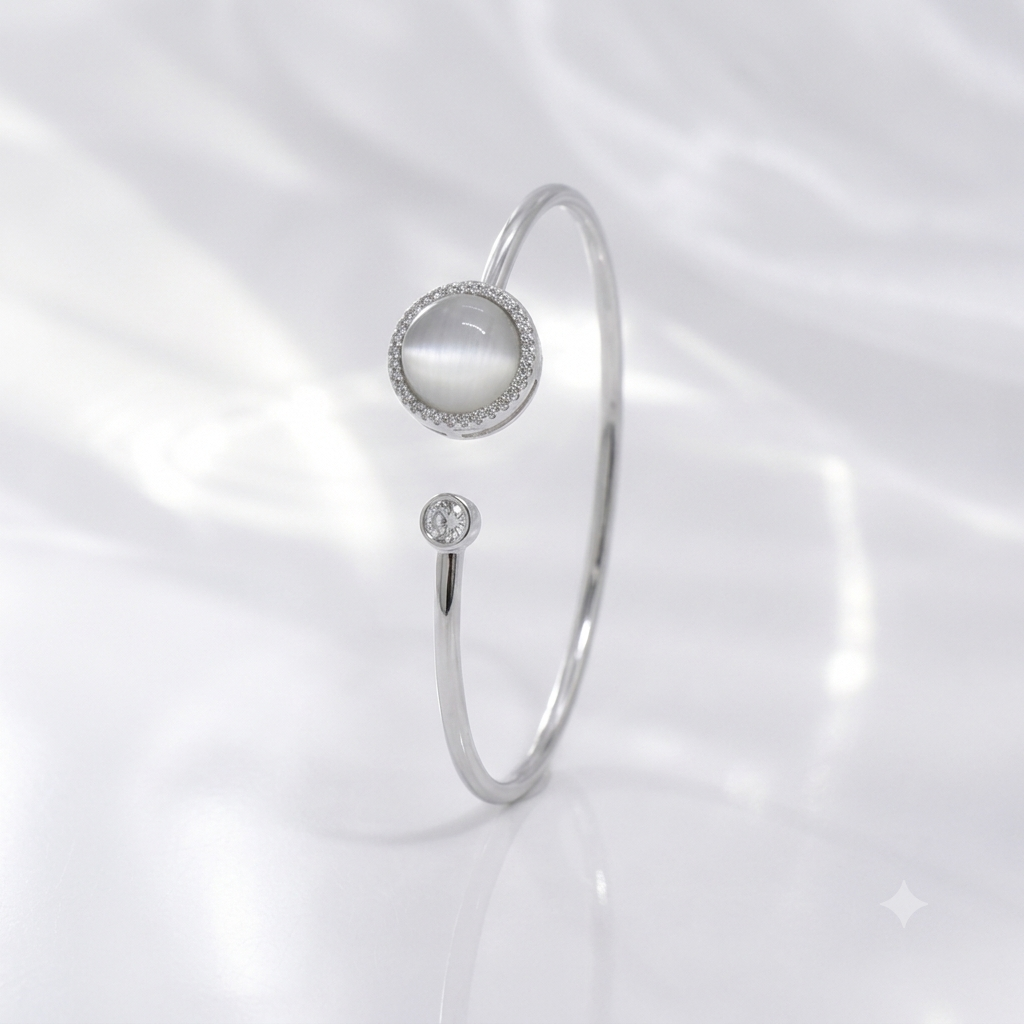A silver-toned open bangle ring featuring a round white mother-of-pearl cabochon surrounded by a halo of small clear stones, with a smaller single clear stone accent below, set against a soft white draped fabric background.